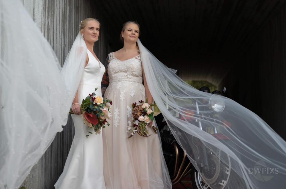 Två kvinnor i eleganta bröllopsklänningar poserar och håller fina buketter